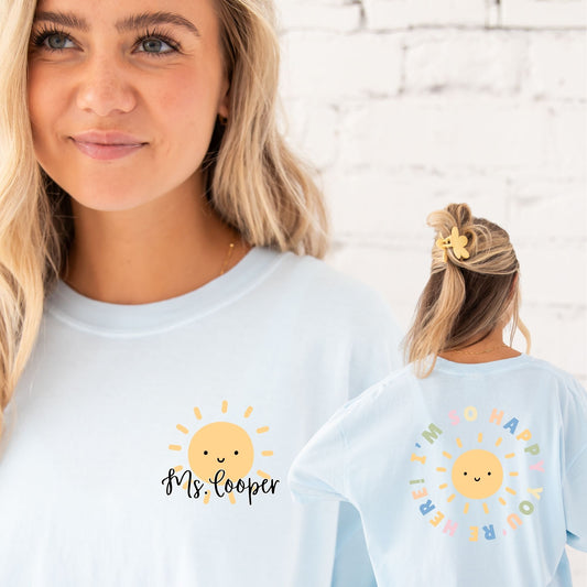 Girl standing in front of a white brick wall wearing an oversized chambray comfort colors t-shirt. the front features a left chest print with a cute smiling yellow sun and a custom script name in black ink. on the back is a large round print with text in multiple colors saying "I'm glad you're here" surrounding a smiling yellow sun