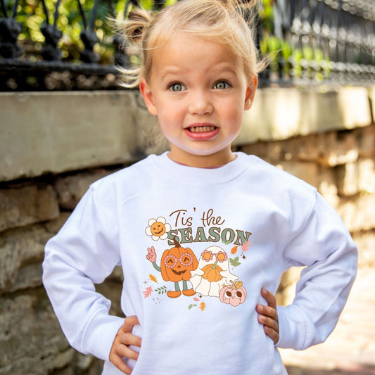 toddler girl wearing a white sweatshirt with a fun and cute halloween print featuring a pumpkin wearing flower glasses and doing a peace sign and a cute ghost wearing flower glasses and holding a flower. Also has falling leaves and a mini pumpkin with a smiley face