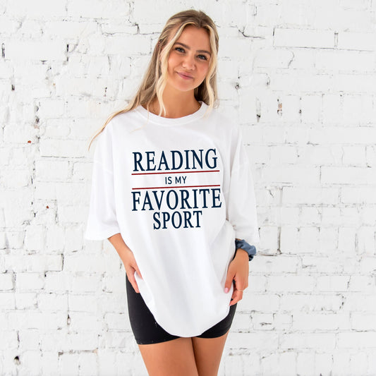 woman standing in front of a white brick wall wearing a white comfort colors t-shirt with a reading is my favorite sport printed design in navy and red ink