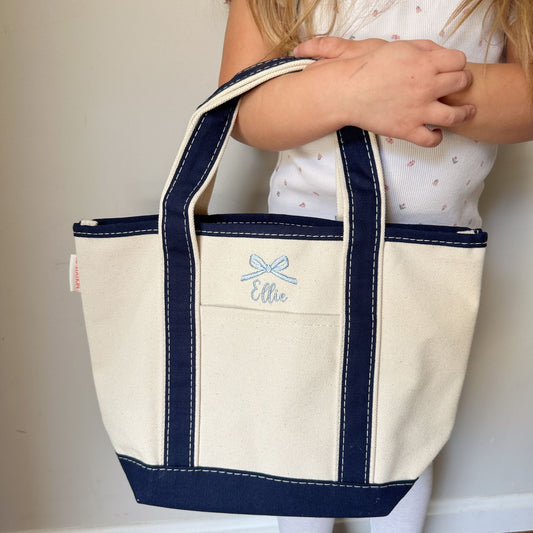 little girl holding a small open top mini canvas tote bag with navy trim and and custom name and mini bow embroidery in powder blue thread