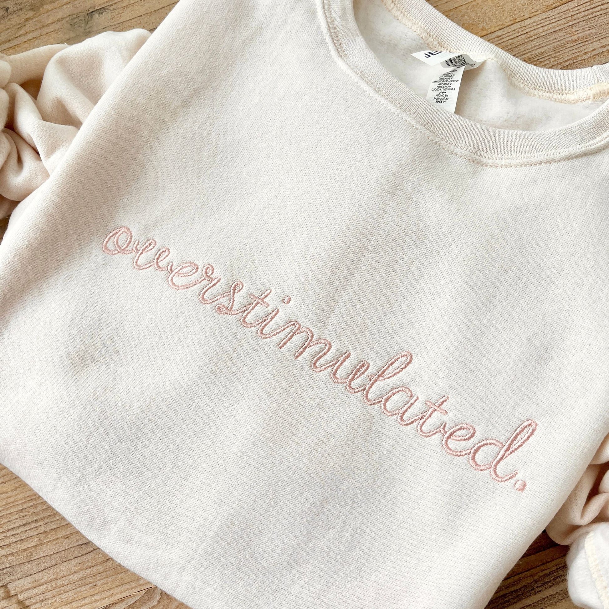 a sweet cream crewneck sweatshirt with custom overstimulated embroidered design in a script font and powder pink thread.
