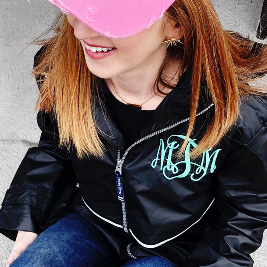 woman sitting outside  wearing a black hooded full zip rain coat with custom monogram embroidered on the left chest in aqua thread.