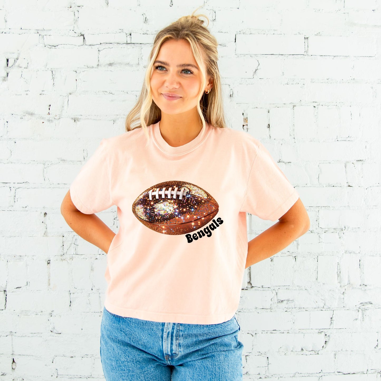 young woman wearing a cropped peachy comfort colors t-shirt with large glitter football design across the chest and bengals text curved under the football in black ink