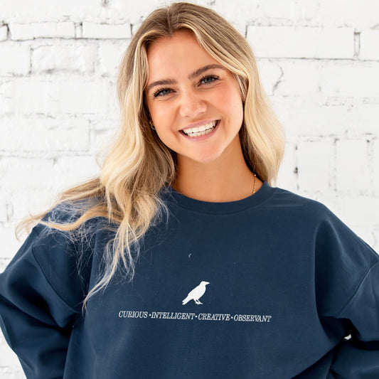 girl wearing navy crewneck sweatshirt with a custom ravenclaw embroidered design featuring a raven and these characteristics: curious, intelligent, creative, observant