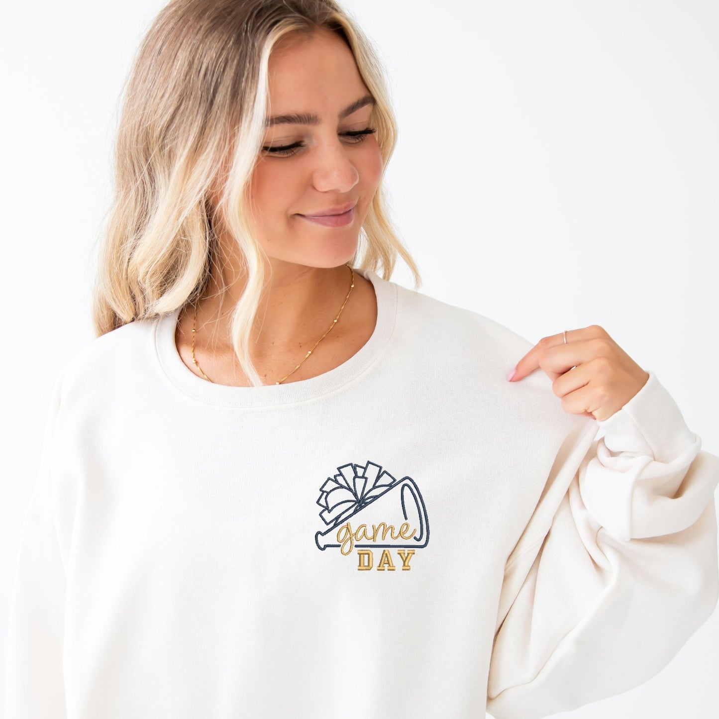 young woman wearing a sweet cream crewneck with ewneck sweatshirt with embroidered cheer megaphone and pom pom with text game day on the left chest innavy and gold threads.