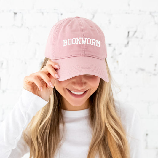 blonde girl wearing a dusty rose baseball hat with bookworm embroidered in athletic font and white thread