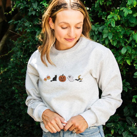 woman wearing a heather oatmeal crewneck sweatshirt with halloween icons embroidered as following: candy corn, black cat, jack-o-lantern, ghost, and bats flying over the moon.