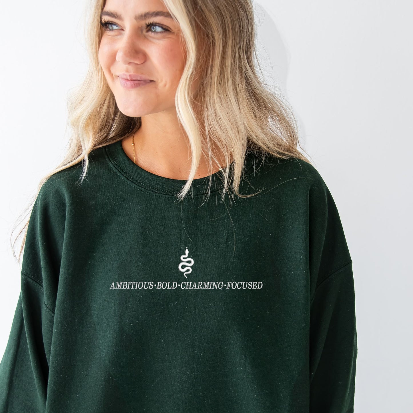 Girl wearing a forest crewneck sweatshirt with custom embroidered Slytherin snake and text reading ambitious, bold, charming, focused.