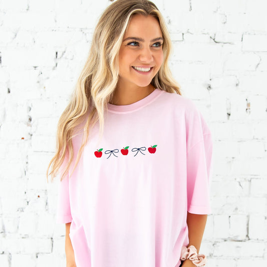 blonde woman wearing an oversized blossom comfort colors t-shirt with an embroidered design featuring alternating mini apples and dainty bows in a navy blue thread. She's standing in front of a white brick wall and has paired the t-shirt with a pair of jeans