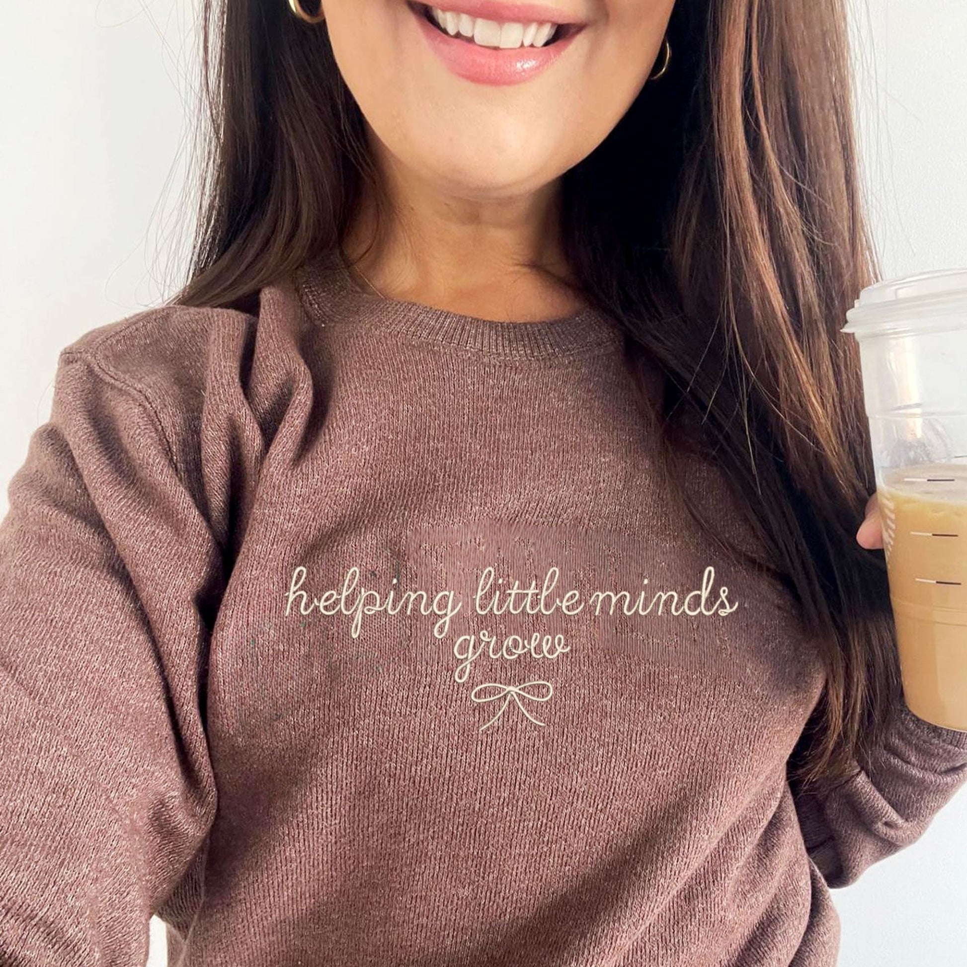 brunette woman holding an iced coffee wearing a terra brown cozy knit nantucket fleece sweatshirt. The sweatshirt features a cute embroidered helping little minds grow design in a script font with a small delicate bow underneath. Embroidery is in camel thread.