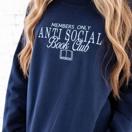 Close up of a girl with long blonde hair wearing a navy crewneck sweatshirt with an embroidered members only antisocial book club design embroidered on the center chest in powder blue thread.