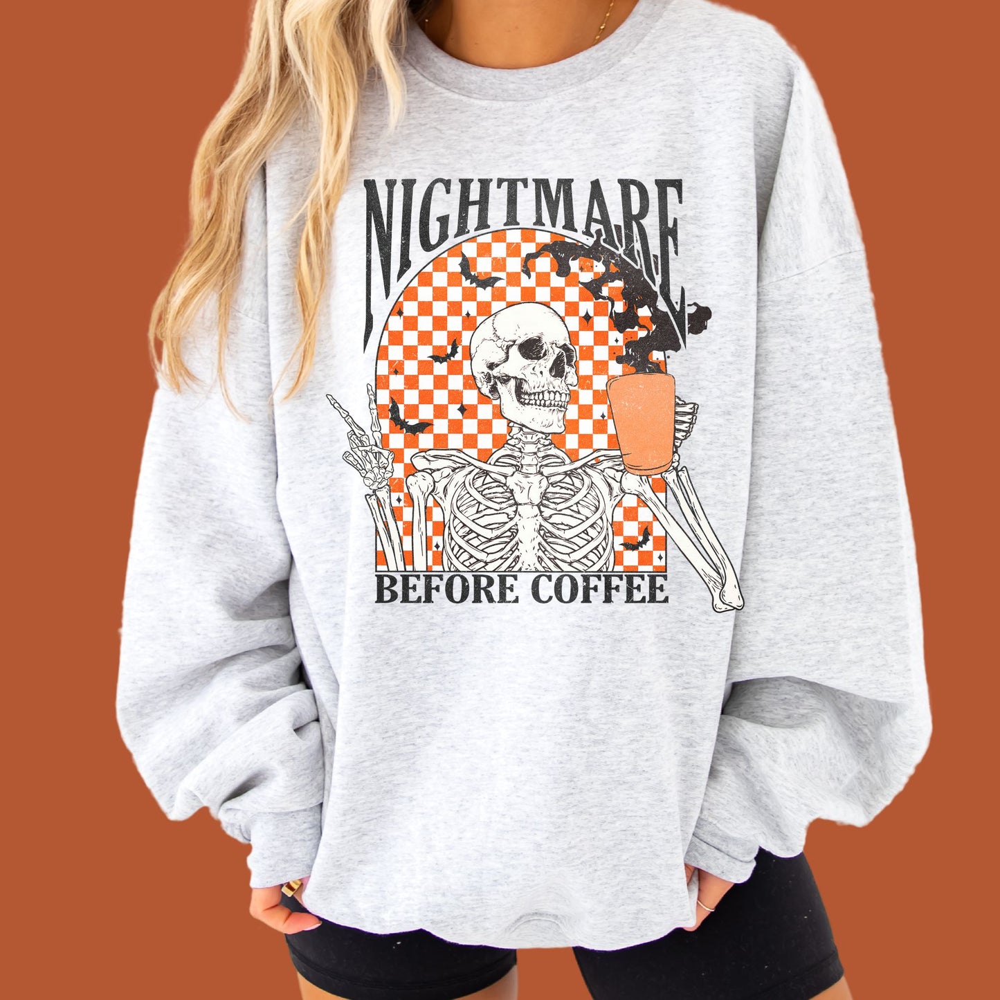 girl wearing biker shorts and an oversized ash crewneck sweatshirt with a funny, vintage, distressed skeleton drinking coffee with the text nightmare before coffee print