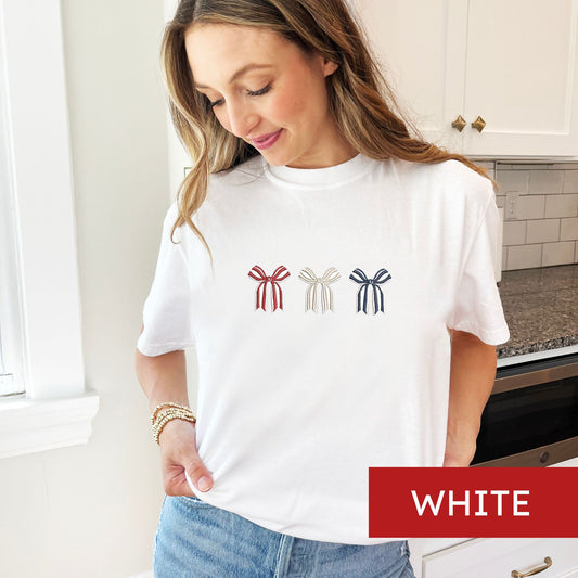 woman wearing a white comfort colors tshirt with embroidered 3 btriped bows in red, white, and navy threads across the chest