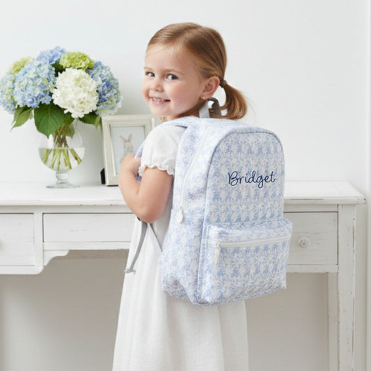 Young girl wearing a blue floral backpack with 'Bridget' embroidered on it, standing in a room with a white desk and flowers.
