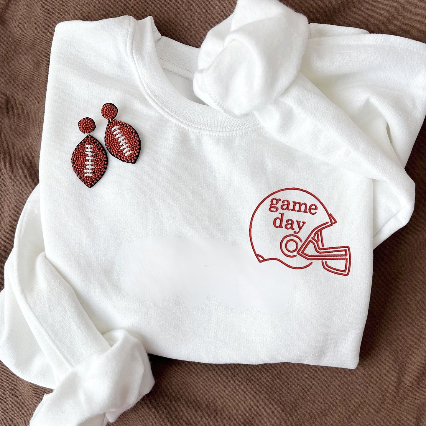 flat lay of a white crewneck sweatshirt with embroidered football helmet and game day text in red on the left chest
