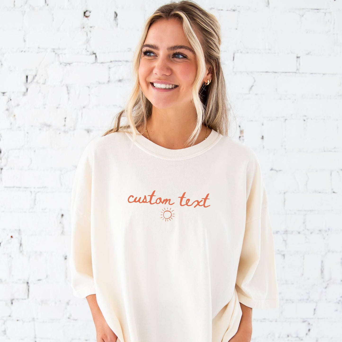girl standing in front of a white brick wall wearing an oversized ivory short sleeve comfort colors t-shirt with personalized custom handwritten script text and sun printed design on the center chest in a soft peach ink.