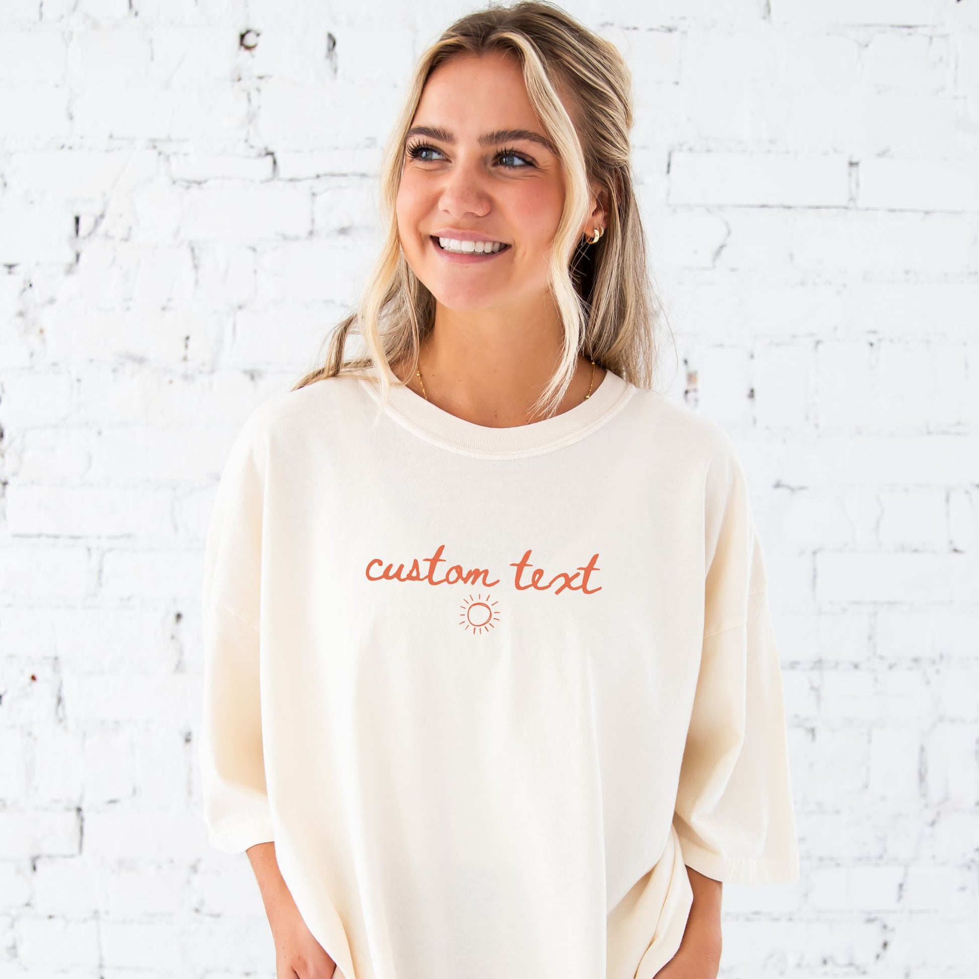 girl standing in front of a white brick wall wearing an oversized ivory short sleeve comfort colors t-shirt with personalized custom handwritten script text and sun printed design on the center chest in a soft peach ink.