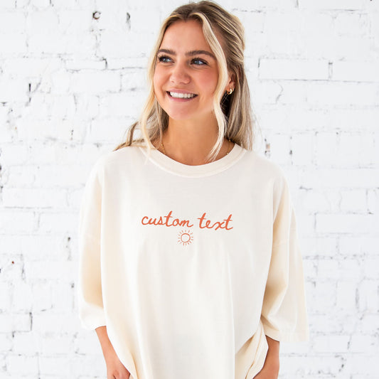 girl standing in front of a white brick wall wearing an oversized ivory short sleeve comfort colors t-shirt with personalized custom handwritten script text and sun printed design on the center chest in a soft peach ink.