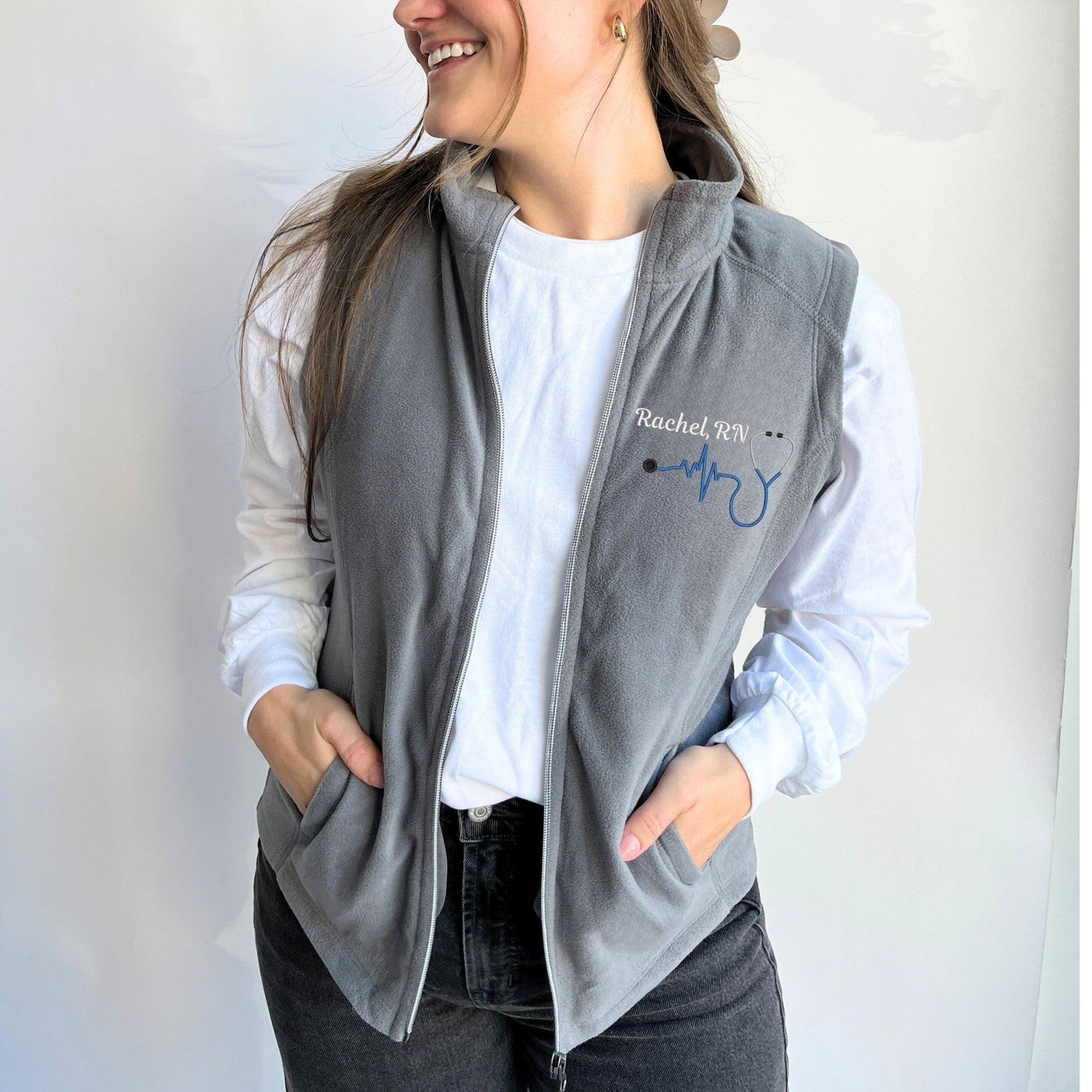 young woman wearing a light weight grey fleece vest with embroidered nurse heartbeat stethoscope in royal thread  on the left chest with name and credentials in white thread.