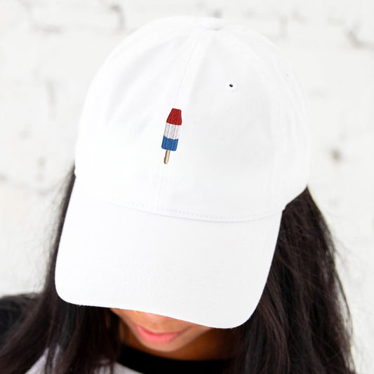 girl with dark hair wearing a white baseball hat with red, white, and blue popsicle embroidered design