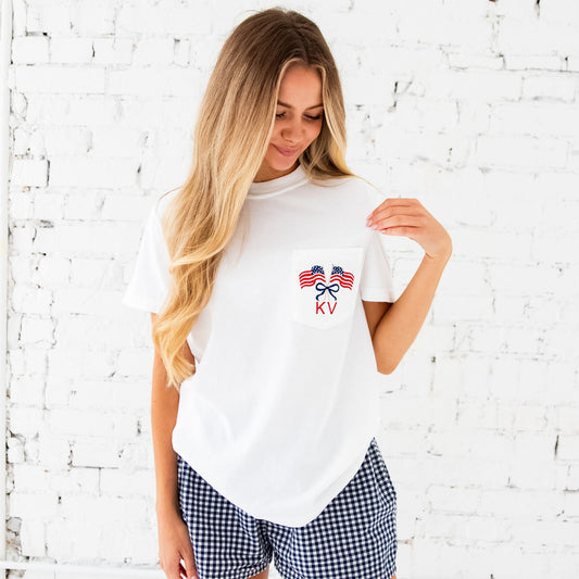 Woman standing in front of a white brick wall. She's wearing a white comfort colors pocket tee with a cute crossed American flags and bow monogram embroidered design on the pocket. Thread colors are a classic red and navy with a touch of camel for the flag poles.
