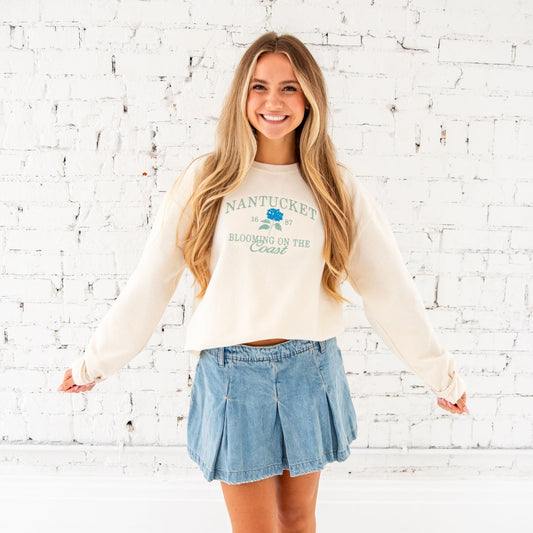 long haired blonde woman standing in front of a white brick wall wearing a sweet cream crewneck sweatshirt with Nantucket 1687 booming on the coast and hydrangea flower embroidered design on the middle chest paired with a denim skirt