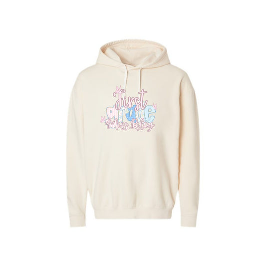 ivory comfort colors hoodie with 'first grade' text in colorful letters on a white background. "First" and the custom name text are in a pink script font, and grade is made out of letters with various patterned letters (gingham, floral, and stripes in pastel colors). The design is also accented by pink butterflies