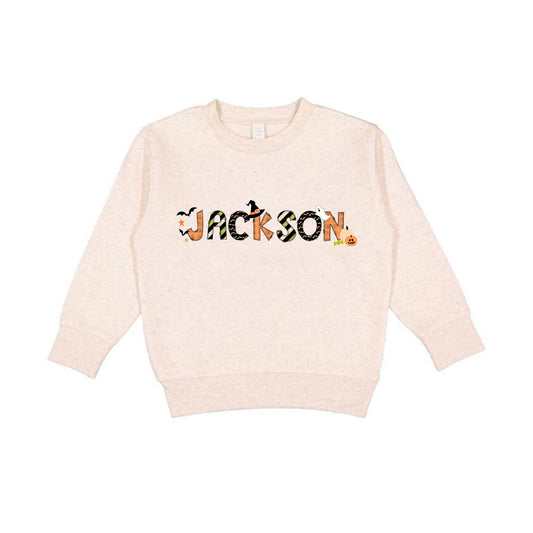 Natural heather crewneck sweatshirt with a custom name print in a mixed patterned Halloween text and accented with cute spooky icons like bats, a ghost, jack-o-lantern, and witch hat.