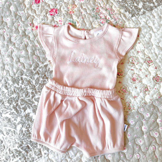 A matching pink set featuring a ruffle short sleeve ribbed bodysuit and pleated shorts. With a custom name embroidered in a cursive script font across the chest in white thread.