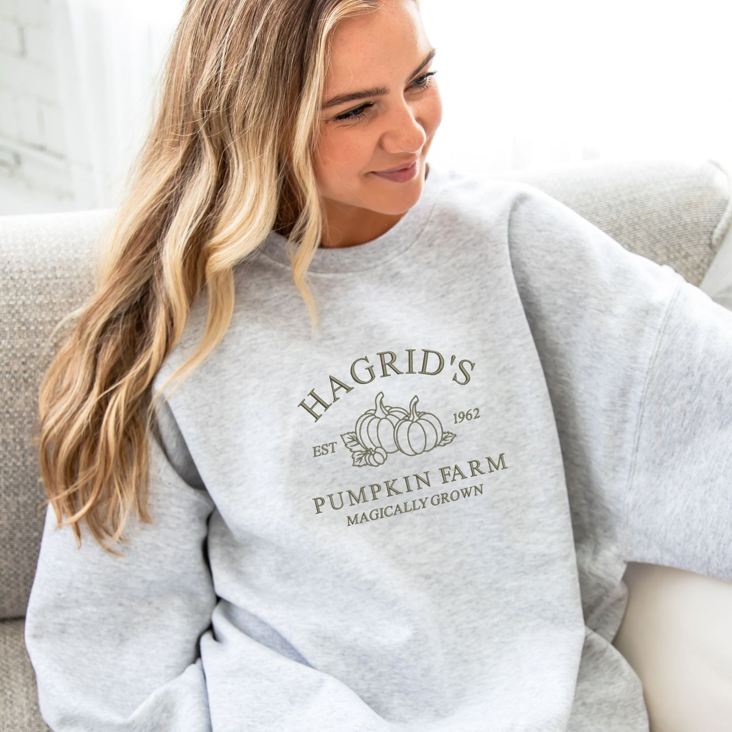 young woman wearing an oversized ash crewneck sweatshirt with embroidered hagrid's pumpkin farm harry potter inspired design in olive thread across the center chest