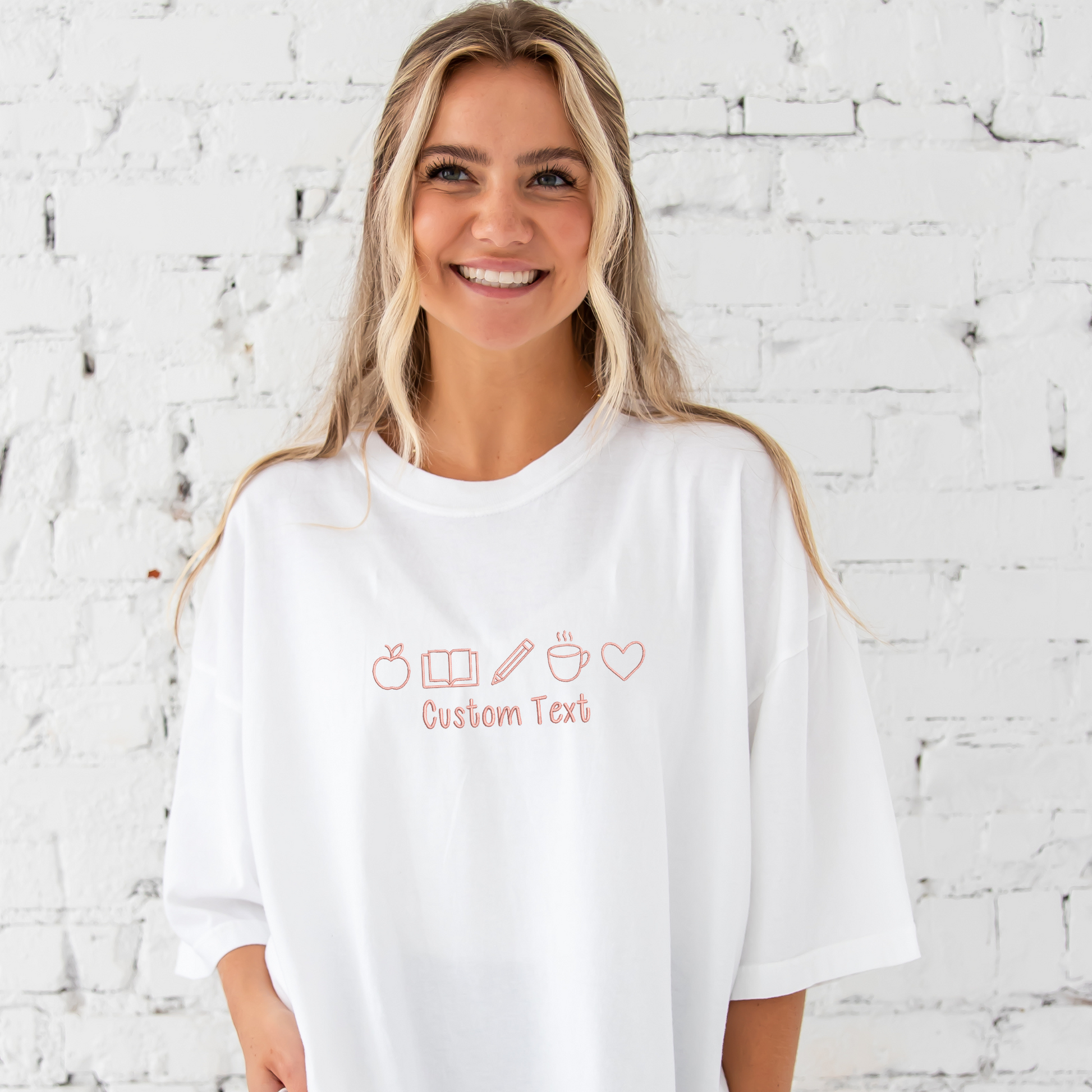 young woman wearing a white comfort colors t-shirt with embroidered teacher icons in coral pink thread across the chest and custom text underneath