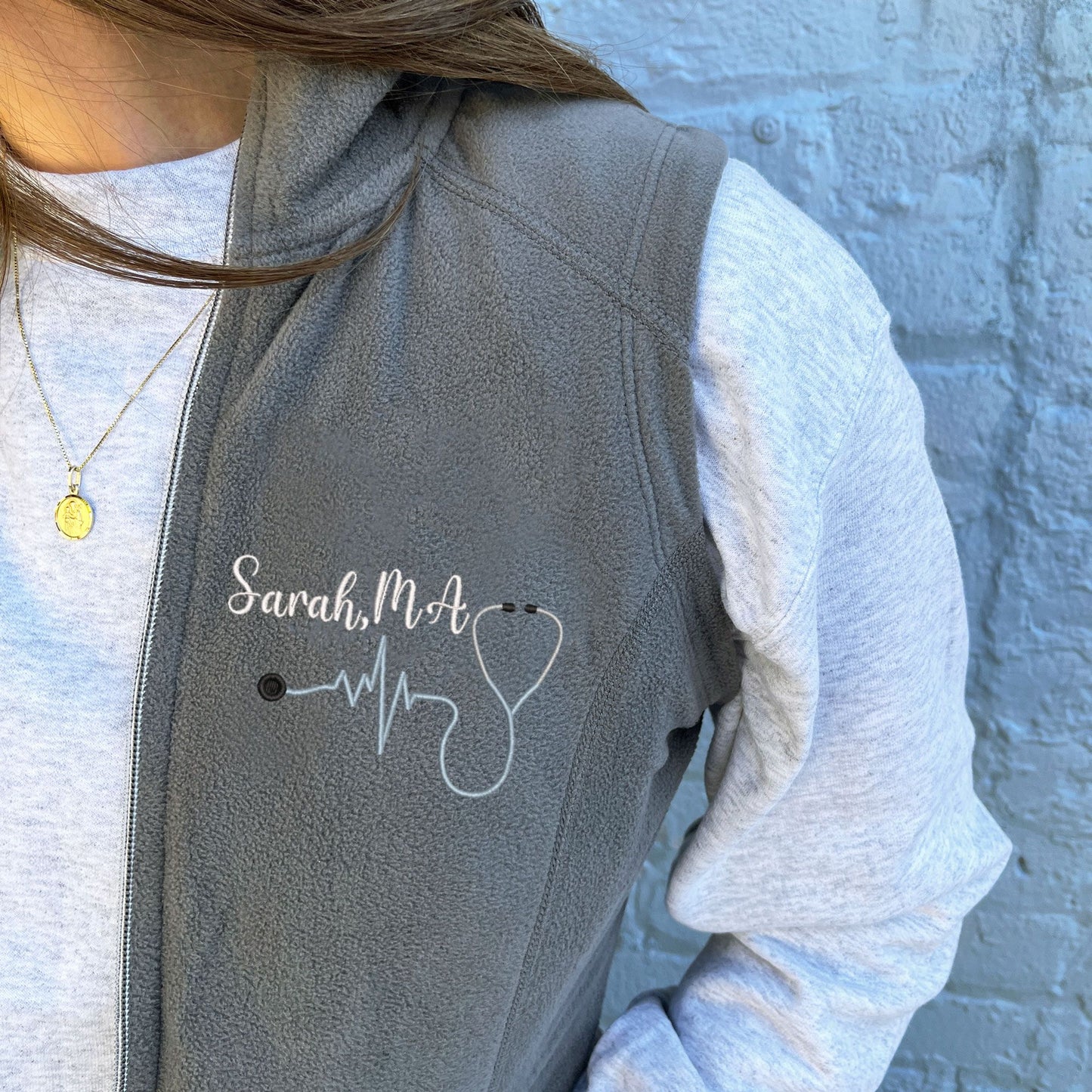 young woman wearing a light weight grey fleece vest with embroidered nurse heartbeat stethoscope in powder blue thread  on the left chest with name and credentials in white thread.