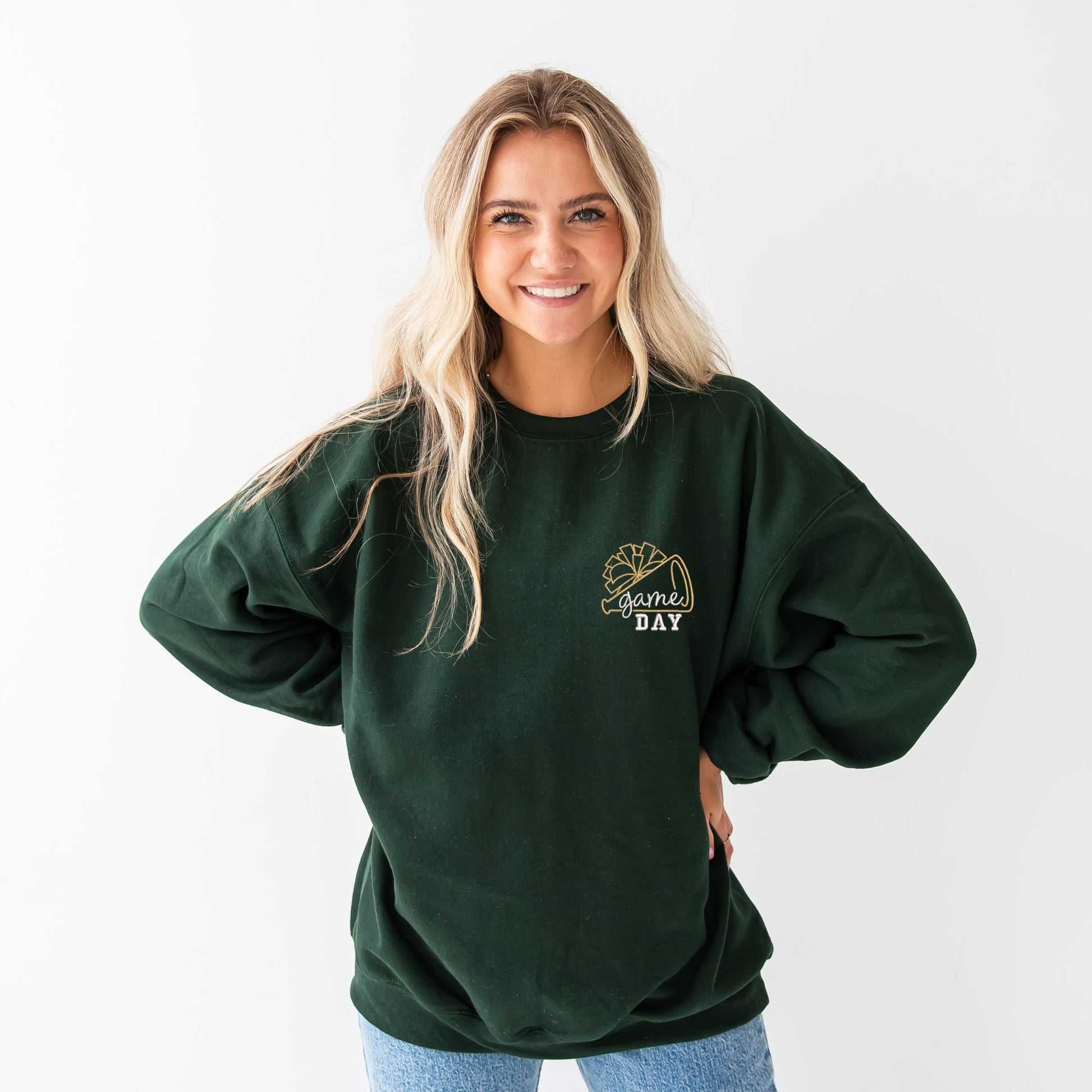 young woman wearing a forest green crewneck sweatshirt with embroidered cheer megaphone and pom pom with text game day on the left chest in gold and white thread