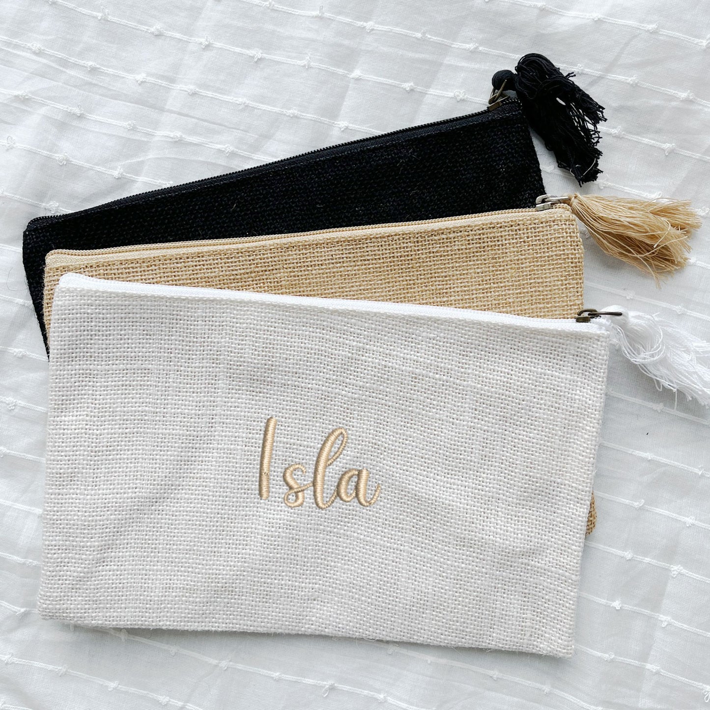 black, natural jute, and white zippered jute pouches with custom name embroidery in a script font and camel thread