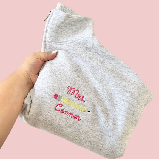 a folded ash quarter zip sweatshirt with a custom curly script name and pencil embroidered design on the left chest