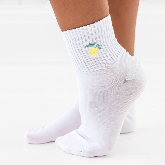 person standing on tip toes, wearing white crew socks featuring a mini lemon embroidered icon.