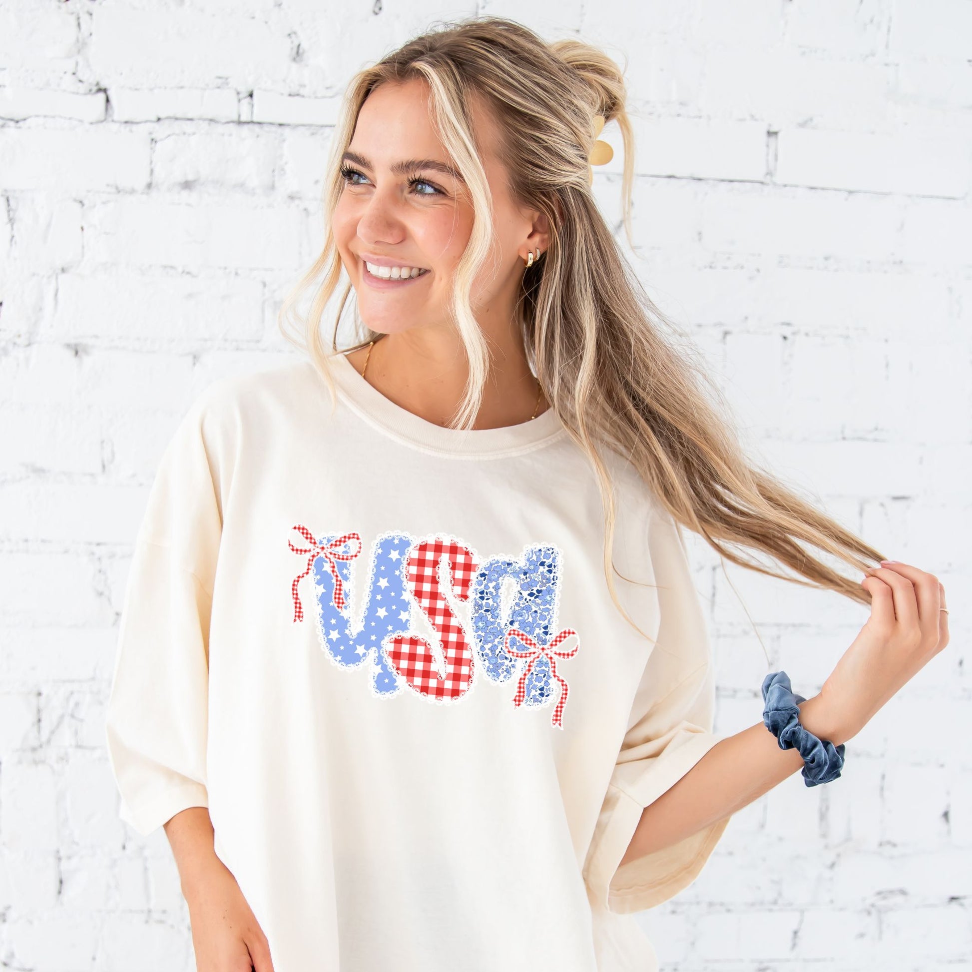 Girl standing in front of a brick wall wearing an ivory comfort colors t-shirt with coquette bow lace usa floral and gingham print on the center chest