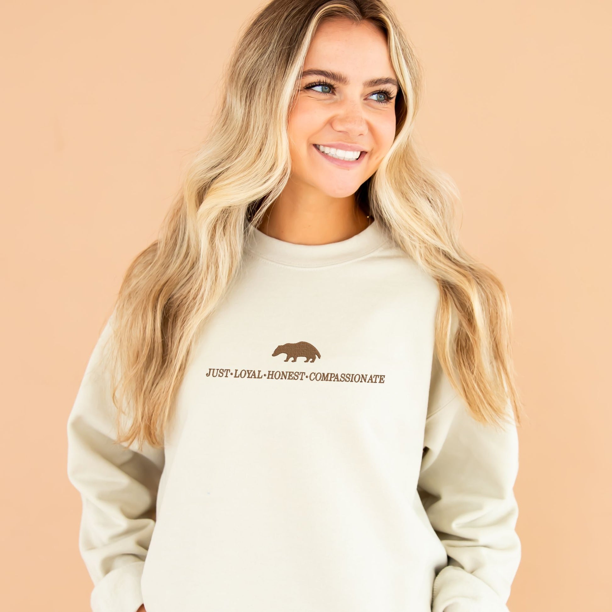 Girl wearing a sand crewneck sweatshirt with custom chestnut embroidered design of a badger and the text: just, loyal, honest, compassionate