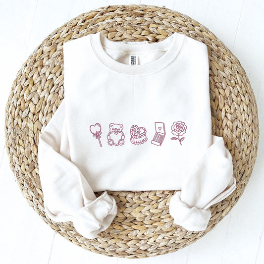 a folded white crewneck sweatshirt with an embroidered design across the chest featuring: heart lollipop, bear with heart, heart cake, matches, and a rose in mauve pink thread