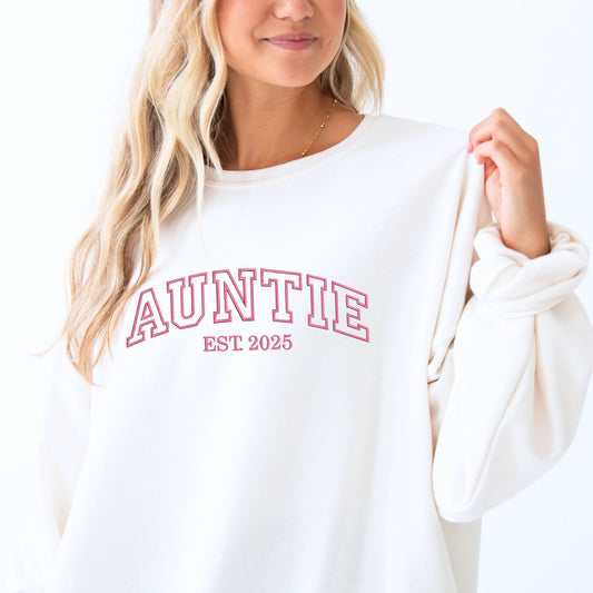 young woman wearing a light blue crewneck sweatshirt with embroidered AUNTIE in all caps athletic block font across the chest  and Est. 2025 underneath in navy thread.