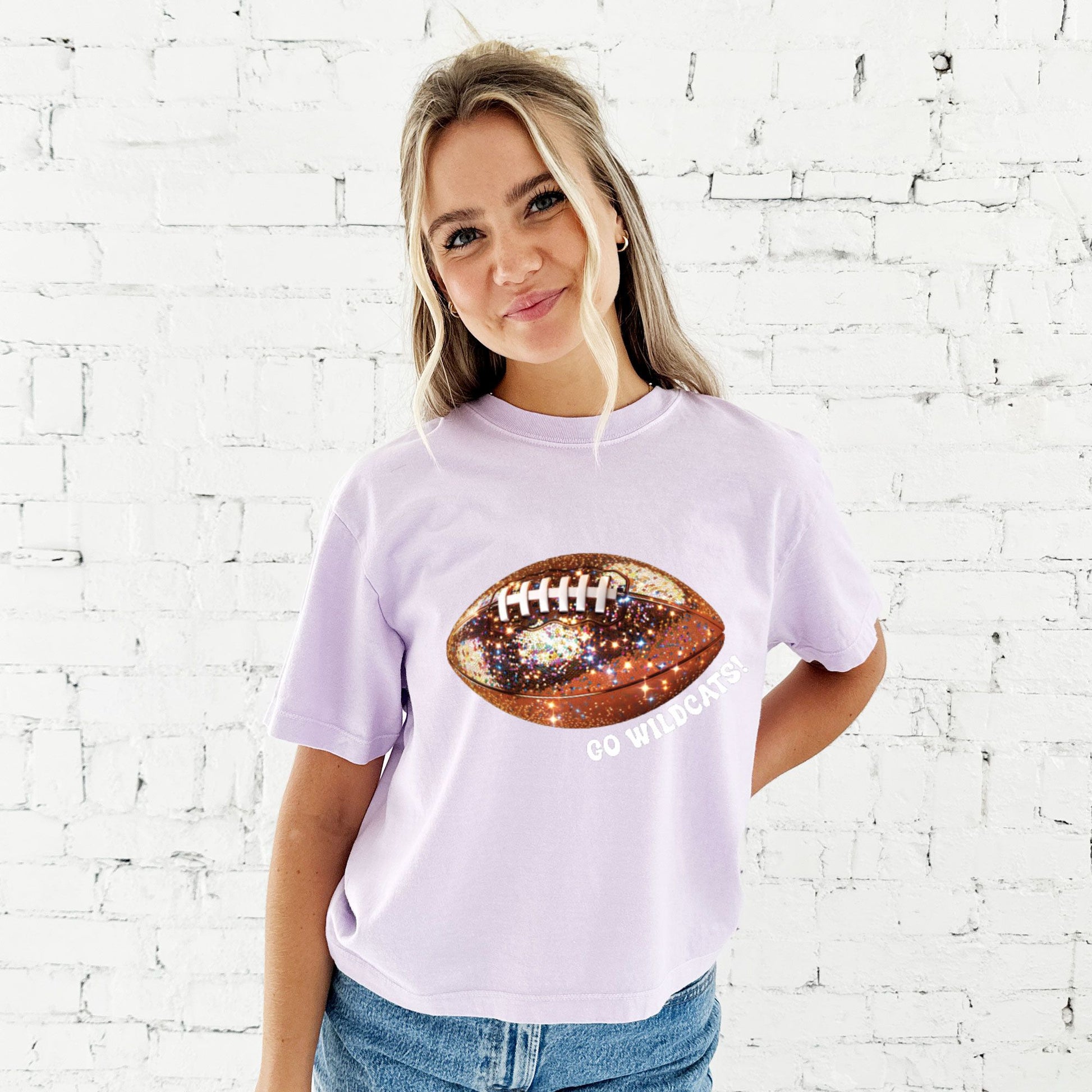 young woman wearing a cropped orchid comfort colors t-shirt with large glitter football design across the chest and go wildcats text curved under the football in white ink