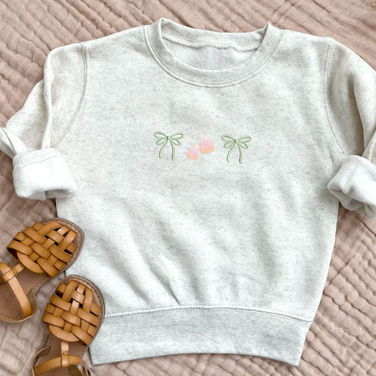 heather oatmeal toddler sweatshirt with custom embroidered double loop bow candy corn bow design across the center chest. Sweatshirt is paired with brown leather sandals and it's laying on a muslin blanket.