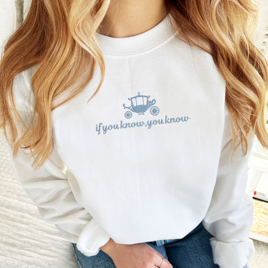 woman wearing an oversized white crewneck sweatshirt with carriage icon and if you know you know embroidered design