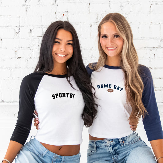 two girls wearing a 3/4 sleeve length black/white and navy/white raglan tee with sports! printed in an arched college block font and black ink on the center chest on one and game day with a mini football printed in navy on the other.