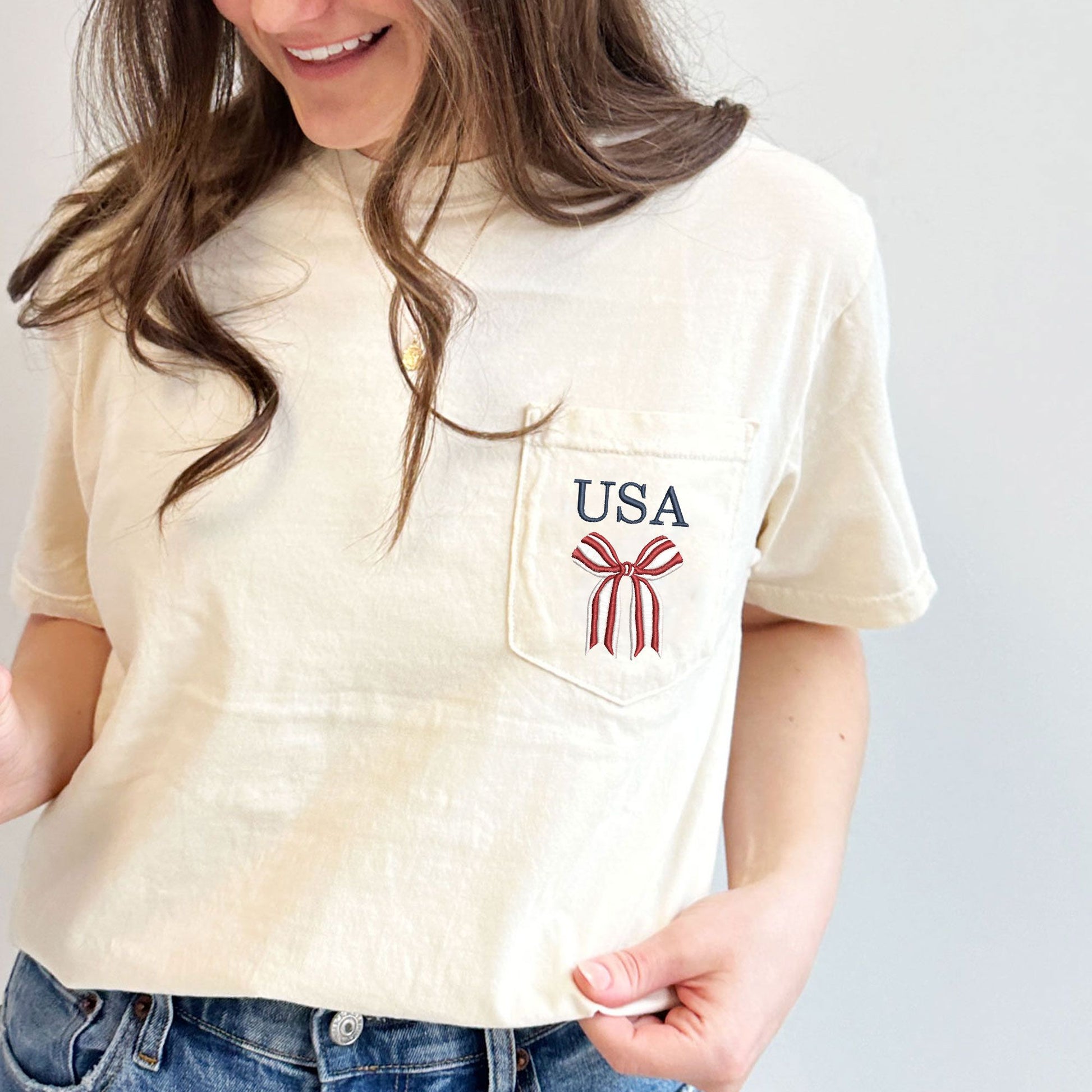woman wearing an ivory  comfort colors pocket tshirt with embroidered red and white striped bow and USA in navy above