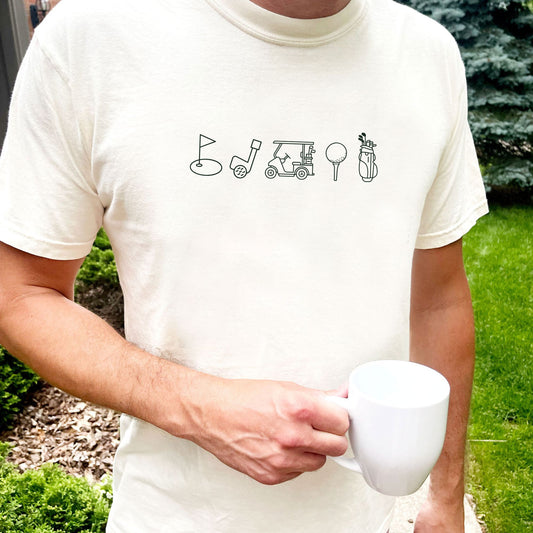 man standing outside wearing an ivory comfort colors t-shirt with custom outlined golf icon printed design featuring: golf bag, golf ball on a tee, golf cart, golf club, and golf green/flag