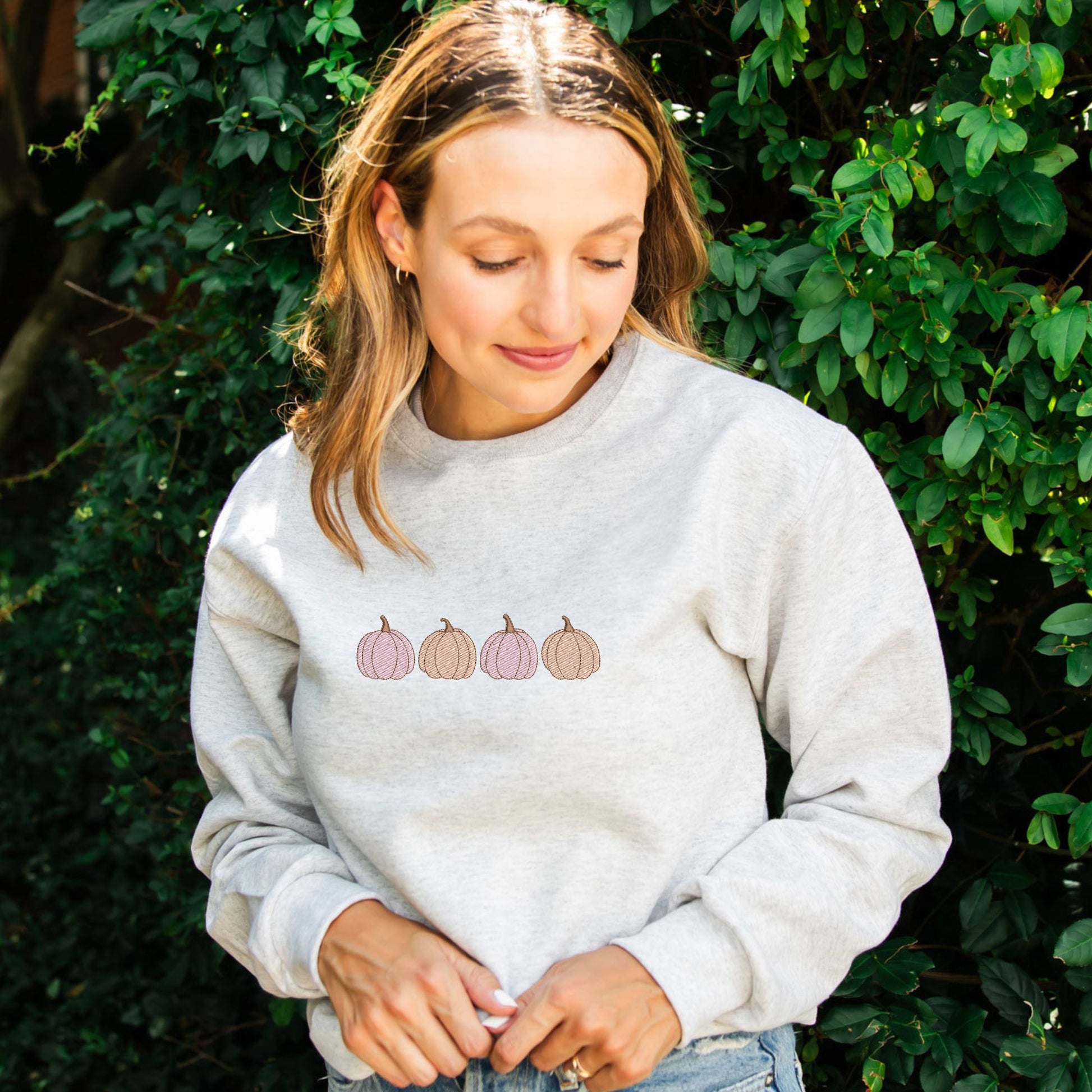 young woman wearing a heather oatmeal crewneck sweatshirt with embroidered 4 pumpkins in a row in alternating pink and orange threads across the chest