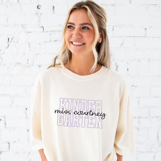 woman wearing an ivory comfort colors t-shirt with a custom grade level in an open block athletic font and custom name in a script font printed on the front center