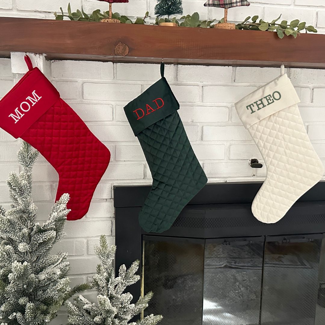three quilted stockings hanging on a mantle in cream, dark green, and red. On the stockings are embroidered name in typewriter font and christmas thread colors.