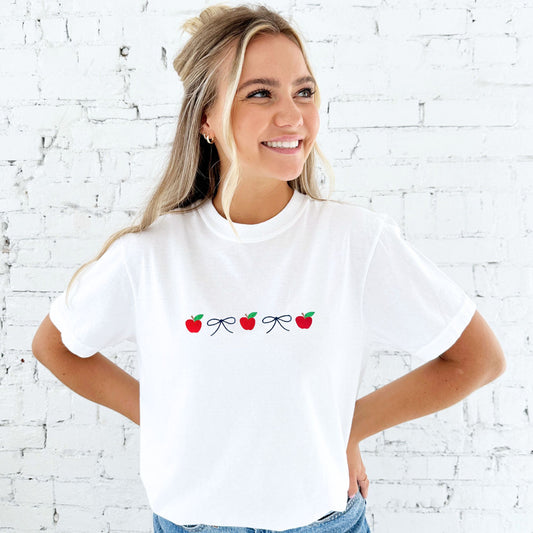blonde woman wearing a white comfort colors t-shirt with an embroidered design featuring alternating mini apples and dainty bows in a navy blue thread. She's standing in front of a white brick wall and has paired the t-shirt with a pair of jeans